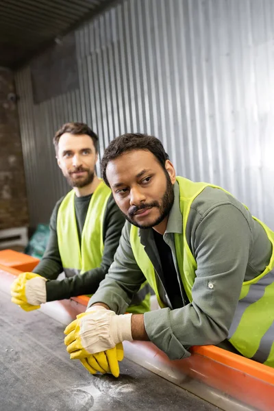 Indian sorter in protective vest and gloves looking away near conveyor and blurred colleague while working together in garbage sorting center, garbage sorting and recycling concept — Stock Photo