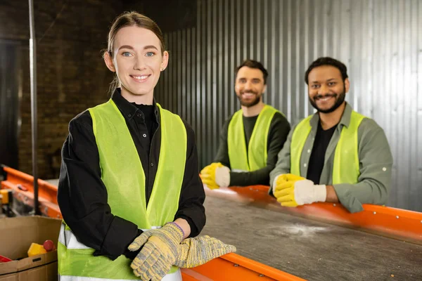 Smiling sorter in protective clothes and gloves looking at camera while standing near conveyor and blurred multiethnic colleagues in garbage sorting center, garbage sorting and recycling concept — Stock Photo