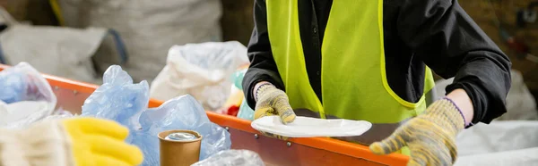 Cropped view of sorter in protective vest and gloves taking garbage from conveyor while working in waste disposal station at background, garbage sorting and recycling concept, banner — Stock Photo