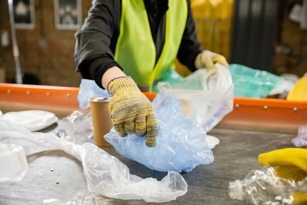 Обрезанный вид размытого работника в защитном жилете и перчатках, забирающего пластиковый пакет с конвейера во время работы на станции утилизации отходов на заднем плане, концепция сортировки мусора — стоковое фото