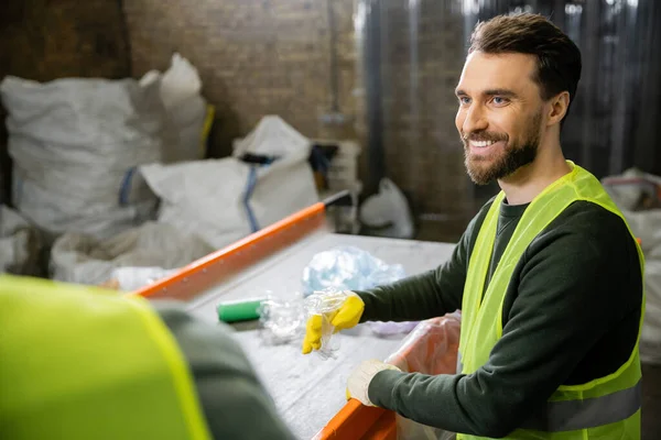 Cheerful male sorter in protective vest and gloves looking at blurred colleague while sorting garbage near conveyor in waste disposal station, garbage sorting and recycling concept — Stock Photo