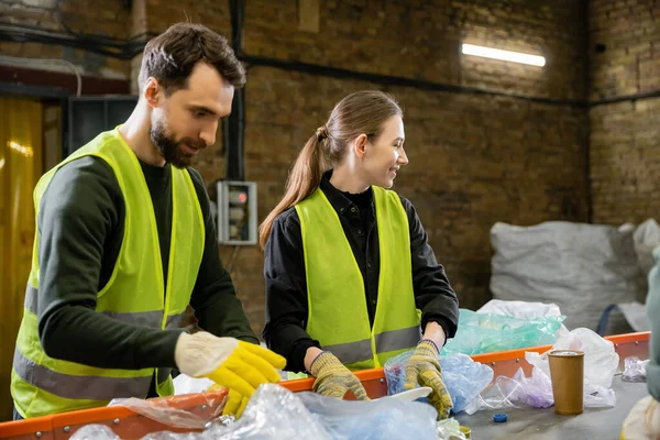 Mujer joven sonriente en ropa protectora y guantes clasificando la basura en el transportador cerca de un colega que trabaja en la estación de eliminación de residuos, clasificación de basura y el concepto de reciclaje - foto de stock