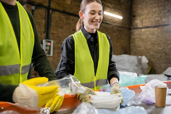 Positiver junger Arbeiter in Handschuhen und Schutzweste, der Plastikmüll in der Nähe von verschwommenem Kollegen und Förderband in Entsorgungsstation, Müllsortier- und Recyclingkonzept hält — Stockfoto