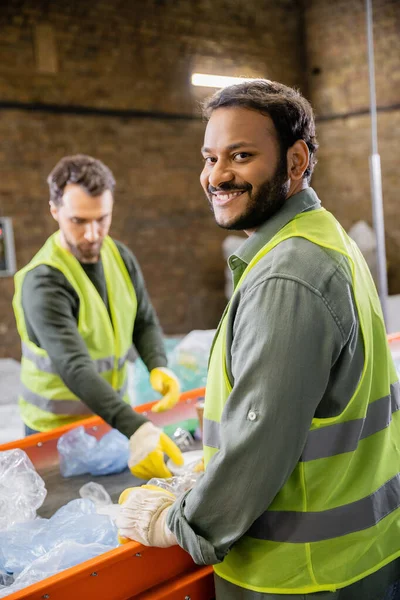 Positive indian worker in protective clothes looking at camera while standing near trash on conveyor and blurred colleague in garbage sorting center, garbage sorting and recycling concept — Stock Photo