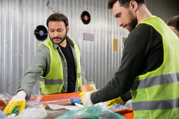 Indian sorter in gloves and protective vest taking trash from conveyor near blurred colleague working together in garbage sorting center, garbage sorting and recycling concept — Stock Photo