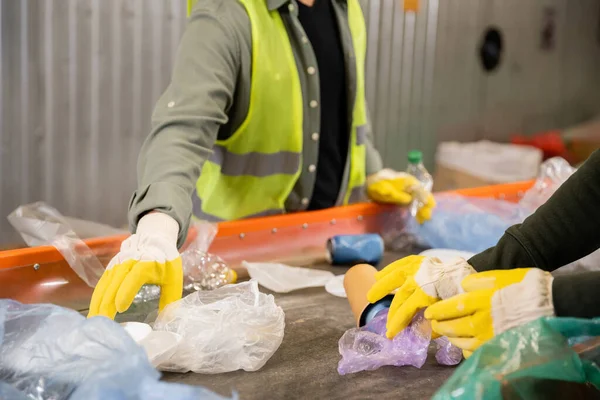 Cropped view of workers in protective gloves taking plastic trash from conveyor while working together in blurred garbage sorting center, garbage sorting and recycling concept — Stock Photo