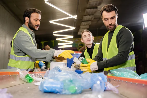 Multiethnic sorters in protective clothes and gloves talking to colleague while working with plastic garbage on conveyor in waste disposal station, garbage sorting and recycling concept — Stock Photo