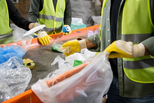 Cropped view of sorter in gloves and protective vest holding blurred plastic bag and taking trash from conveyor while working near colleagues in waste disposal station, garbage recycling concept — Stock Photo