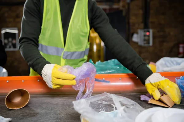 Обрізаний вид сортувальника в захисних рукавичках і жилеті, що приймає сміття з конвеєра, працюючи в центрі сортування розмитих сміття, концепції сортування сміття та переробки — стокове фото
