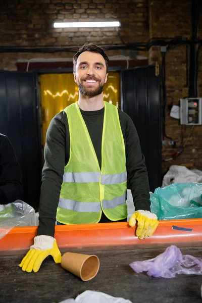 Smiling and bearded sorter in gloves and protective vest looking at camera while working with trash on conveyor in garbage sorting center, garbage sorting and recycling concept — Stock Photo