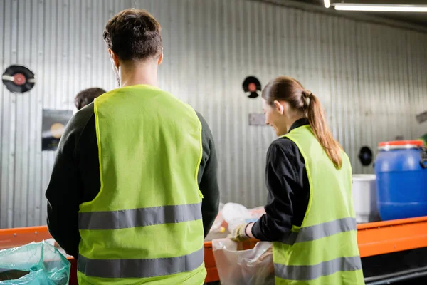 Arbeiter in Warnweste in Uniform trennt Müll in der Nähe verschwommener Kollegen und Förderer im Müllsortierzentrum im Hintergrund, Müllsortier- und Recyclingkonzept — Stockfoto