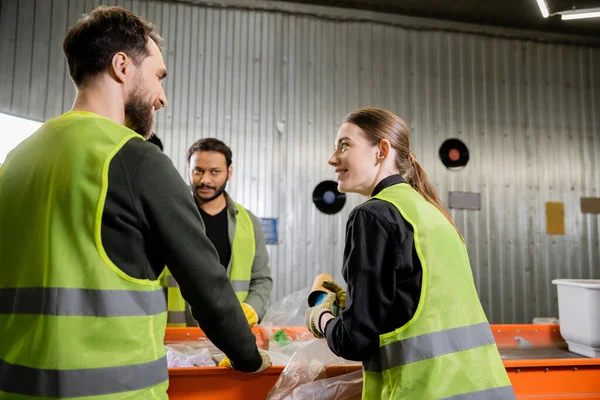Lächelnde Frau in Schutzuniform und Handschuhen, die Müll in der Hand hält und mit Kollegen spricht, während sie in der Müllentsorgungsstation in der Nähe von Förderbändern zusammenarbeitet. — Stockfoto