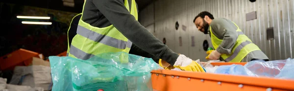 Male worker in protective gloves and reflective vest taking garbage from conveyor near plastic bag and blurred indian colleague at background in waste disposal station, recycling concept, banner — Stock Photo