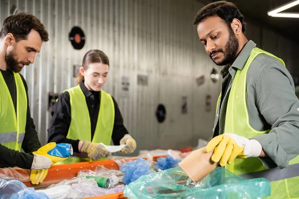 Bearded indian worker in safety vest and protective gloves putting paper cup in plastic bag while separating trash near conveyor and colleagues in waste disposal station, recycling concept — Stock Photo