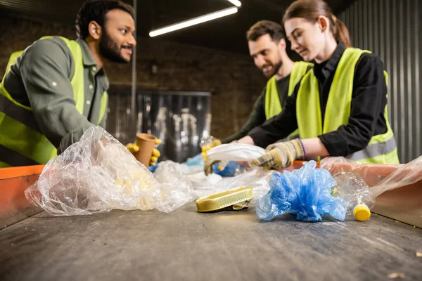 Lixo plástico em transportador e trabalhadores multiétnicos desfocados em coletes de segurança e luvas trabalhando juntos em segundo plano na estação de eliminação de resíduos, conceito de reciclagem — Fotografia de Stock