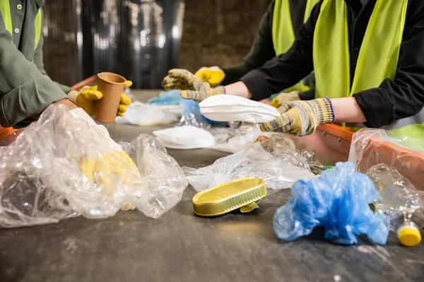 Cropped view of workers in high visibility vests and gloves separating different plastic and paper trash on conveyor while working in waste disposal station, recycling concept — Stock Photo