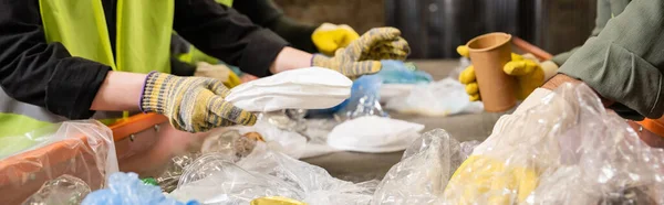Cropped view of workers in protective gloves taking different plastic and paper trash from conveyor while working together in waste disposal station, recycling concept, banner — Stock Photo