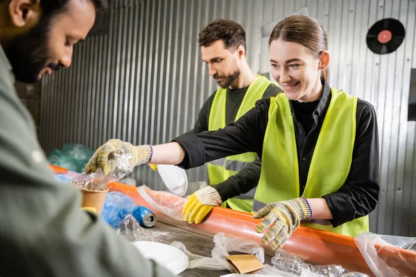 Positive worker in protective gloves and reflective vest taking plastic trash from conveyor near blurred interracial colleagues working in garbage sorting center, recycling concept — Stock Photo