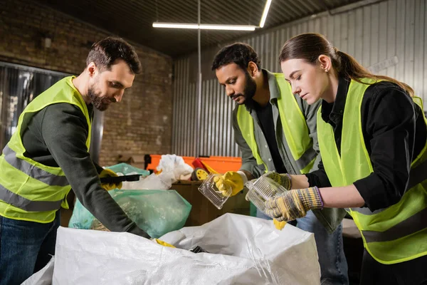 Interracial sorters in protective gloves and safety vests taking plastic containers from sacks while sorting trash together in waste disposal station, garbage sorting and recycling concept — Stock Photo