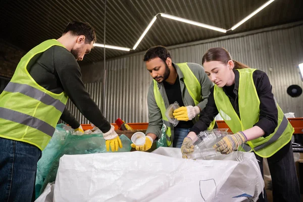 Young worker in reflective vest and gloves holding plastic containers near sack while sorting trash with multiethnic colleagues in waste disposal station, garbage sorting and recycling concept — Stock Photo