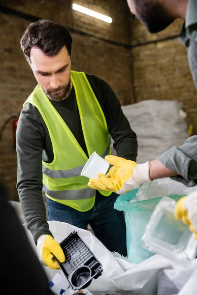 Uomo barbuto in giubbotto riflettente e guanti che tengono rifiuti di plastica vicino al sacco e sfocato collega indiano mentre lavorano insieme nella stazione di smaltimento dei rifiuti, concetto di riciclaggio — Foto stock