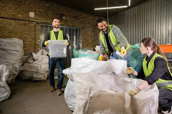 Smiling indian worker in safety vest and gloves giving plastic trash to colleague near sacks and sorter with container while working in waste disposal station, recycling concept — Stock Photo