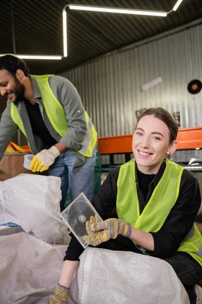 Lächelnder junger Arbeiter in reflektierender Weste und Handschuhen, der in die Kamera blickt, während er Plastikmüll zum Recyceln in der Nähe von Säcken hält und verschwommener indischer Kollege in der Müllentsorgungsstation, Mülltrennung — Stockfoto