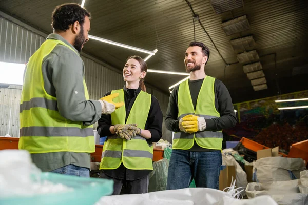 Bearded indian worker in protective glove and vest talking on cheerful colleagues while standing together near blurred sacks with trash in waste disposal station, recycling concept — Stock Photo