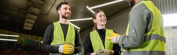 Cheerful workers in protective gloves and high visibility vests looking at indian colleague talking while standing together in blurred waste disposal station, recycling concept, banner — Stock Photo