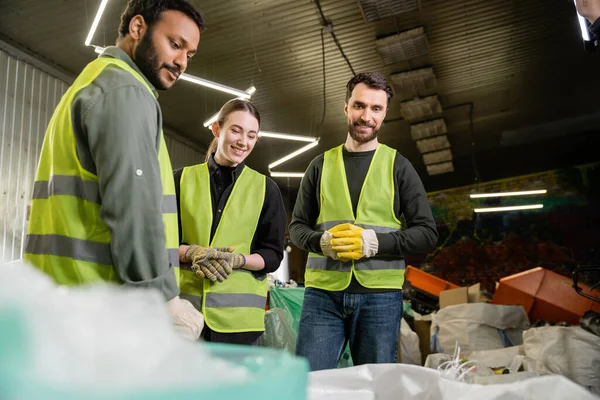 Trabajadores multiétnicos sonrientes con chalecos y guantes protectores que miran sacos borrosos con basura para reciclar mientras trabajan juntos en la estación de eliminación de residuos, concepto de reciclaje - foto de stock