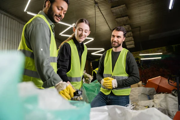 Smiling workers in high visibility vests standing near indian colleague in protective glove holding trash near sacks while working together in blurred waste disposal station, garbage recycling — Stock Photo
