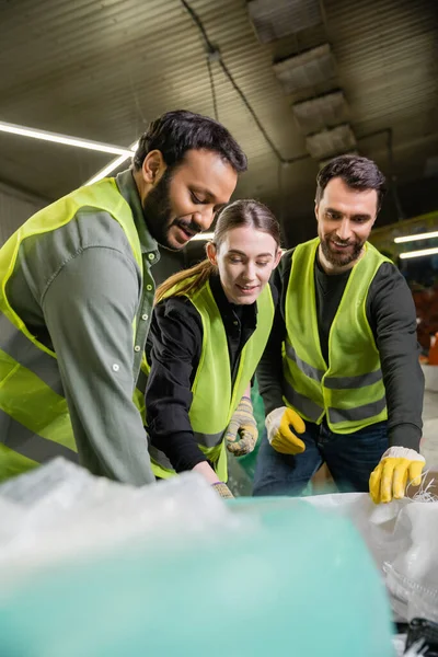 Trabalhadores multiétnicos sorridentes em roupas e luvas de proteção conversando e trabalhando perto de sacos borrados juntos na estação de eliminação de resíduos, processo de triagem de lixo — Fotografia de Stock