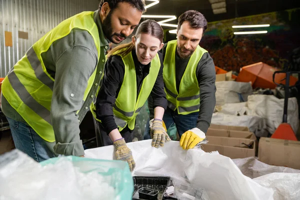 Young female worker in high visibility jacket and gloves looking at sacks while working together with multiethnic male colleagues in blurred waste disposal station, garbage sorting process — Stock Photo