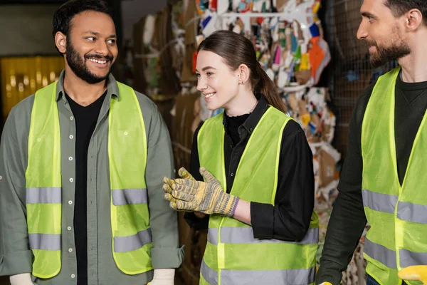 Cheerful female worker in protective vest and gloves looking away while standing near multiethnic colleagues and resting in blurred waste disposal station near waste paper, garbage sorting process — Stock Photo