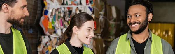 Positive young woman in high visibility vest looking away while standing and resting near interracial colleagues in blurred waste disposal station near waste paper, garbage sorting process, banner — Stock Photo