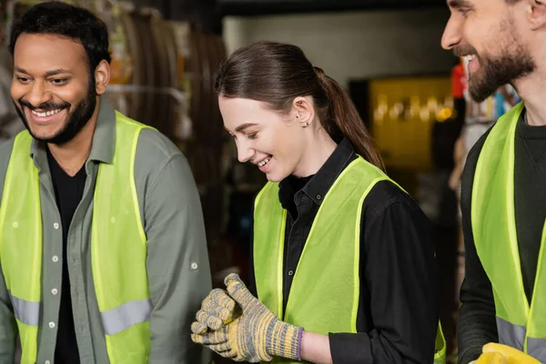 Lächelnde junge Arbeiterinnen in Schutzhandschuhen und Weste, die neben multiethnischen Kollegen stehen, während sie in einer verschwommenen Müllentsorgungsstation zusammenarbeiten. — Stockfoto