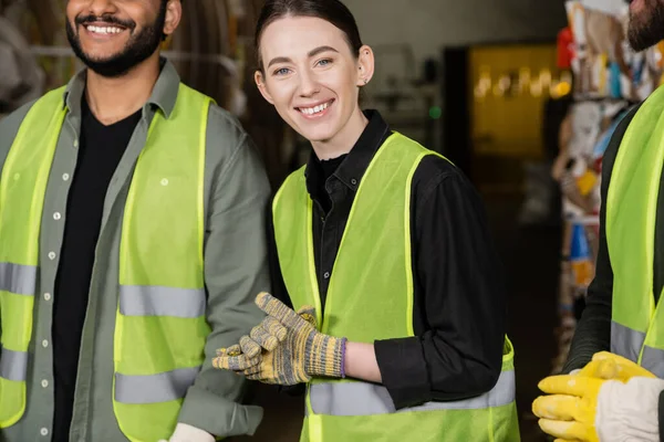 Young worker in high visibility vest and gloves smiling at camera while standing near multiethnic colleagues in blurred waste disposal station, garbage sorting process — Stock Photo