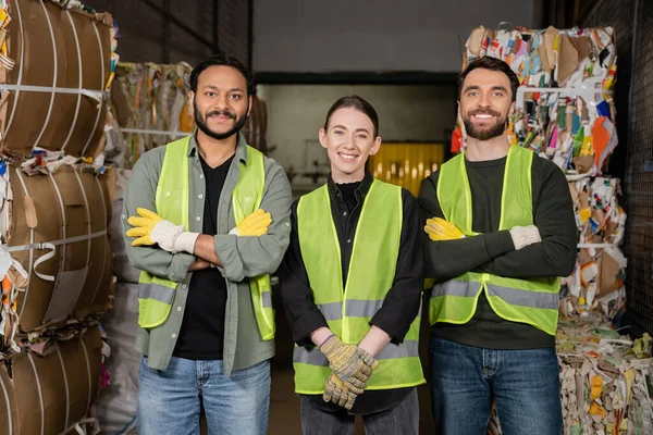 Cheerful multiethnic workers in high visibility vests and gloves crossing arms and looking at camera while standing near waste paper on disposal station, garbage sorting process — Stock Photo