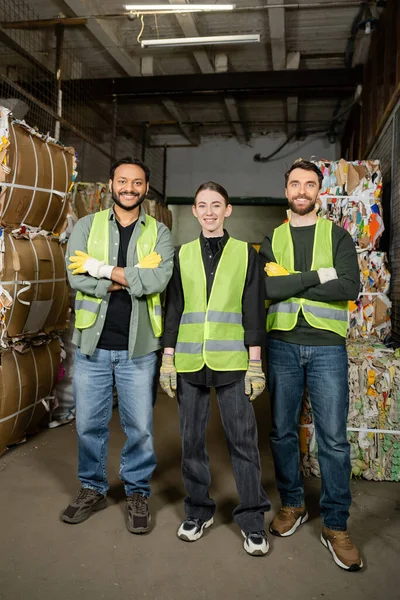 Smiling and interracial workers in high visibility vests and gloves crossing arms and looking at camera while standing near waste paper on disposal station, garbage sorting process — Stock Photo