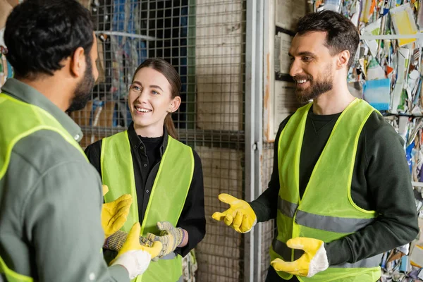 Cheerful man in high visibility vest and gloves talking to multiethnic colleagues while standing and resting in garbage sorting center, waste sorting and recycling concept — Stock Photo