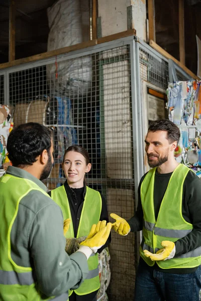 Smiling male worker in safety vest and gloves talking to indian colleague while resting near waste paper in garbage sorting center, waste sorting and recycling concept — Stock Photo