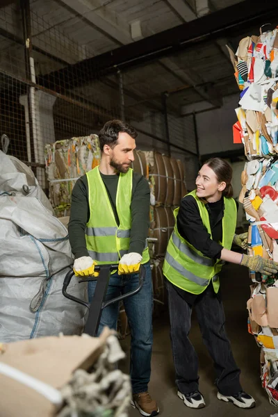Smiling woman in high visibility vest and gloves talking to male colleague with hand pallet truck while working with waste paper in blurred garbage sorting center, waste sorting and recycling concept — Stock Photo