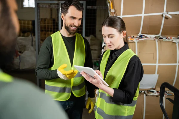 Smiling worker in safety vest and gloves talking to colleague with digital tablet near blurred indian sorter and waste paper in garbage sorting center, waste sorting and recycling concept — Stock Photo