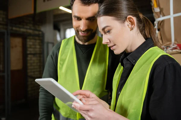 Young worker in safety reflective vest using digital tablet while standing near smiling colleague and blurred waste paper in garbage sorting center, waste sorting and recycling concept — Stock Photo