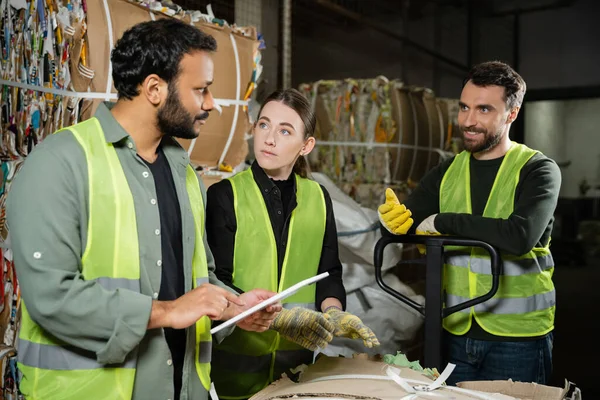 Smiling worker in reflective vest and gloves talking to multiethnic colleagues with digital tablet and standing near waste paper on hand pallet truck in garbage sorting center, waste recycling — Stock Photo