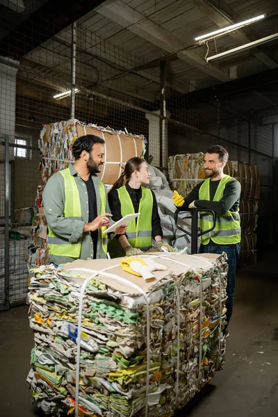 Cheerful sorter in protective vest and gloves talking to interracial colleagues with digital tablet and waste paper on hand pallet truck in waste disposal station, garbage recycling concept — Stock Photo