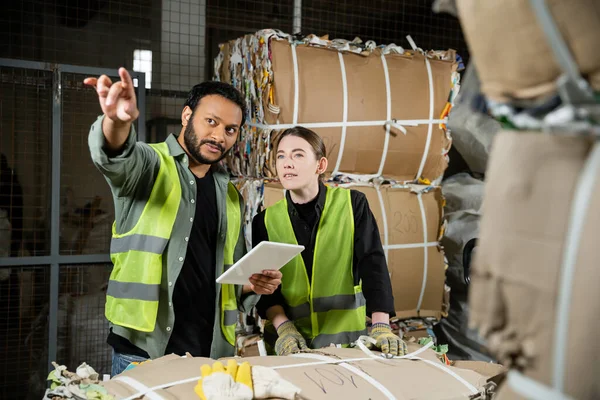 Indian worker in high visibility vest pointing with finger and using digital tablet near female colleague and waste paper on hand pallet truck waste disposal station, garbage recycling concept — Stock Photo