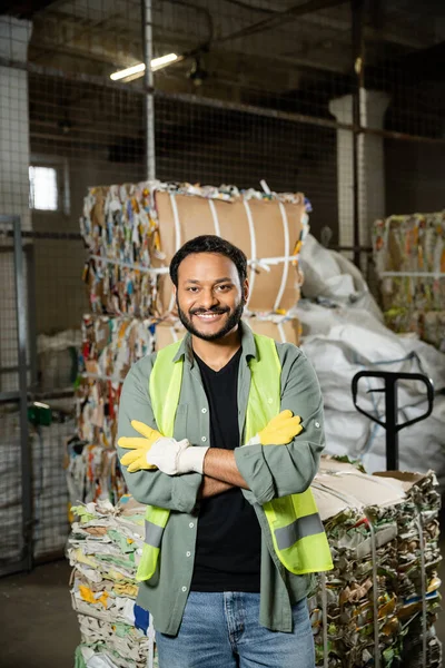 Trabalhador sorridente e barbudo em luvas de proteção e colete cruzando os braços e olhando para a câmera enquanto estava perto de papel residual borrado na estação de eliminação de resíduos, triagem de lixo e conceito de reciclagem — Fotografia de Stock