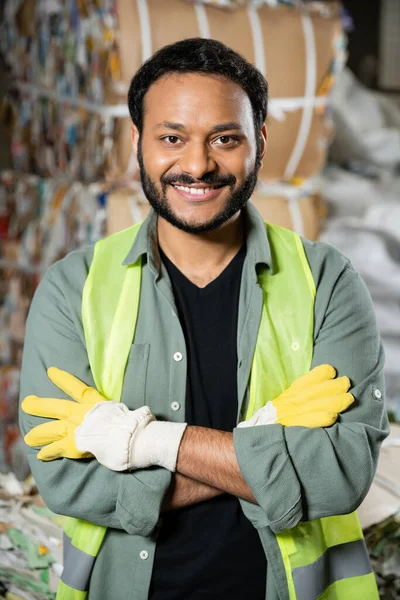 Веселый индийский рабочий в рефлекторном жилете и перчатках смотрит в камеру и скрещивает руки, стоя рядом с бумагой на размытом фоне в мусорной станции, концепция переработки мусора — стоковое фото