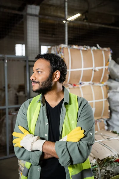 Bearded indian worker in high visibility vest and gloves looking away and crossing arms while standing near blurred waste paper in waste disposal station, garbage sorting and recycling concept — Stock Photo